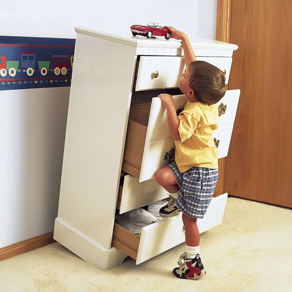 Child climbing on a white dresser with open drawers, demonstrating the risk of furniture tipping without safety straps.