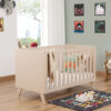 Babymore Dante Cot Bed in natural wood finish styled in a modern nursery with toys and wall art.
