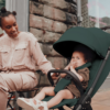 A lifestyle image of a smiling mother interacting with her baby, who is comfortably seated in the deep green stroller. The baby looks playfully at the camera, while the mother, dressed in neutral tones, is seated at an outdoor café with a brick wall in the background.