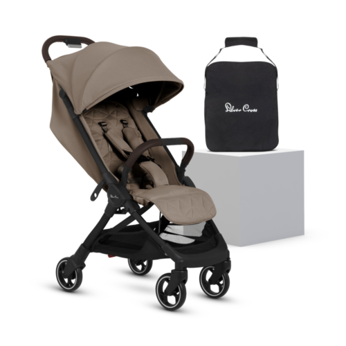 A stylish, compact baby stroller in a neutral beige color with a black frame and four swivel wheels. It has a quilted seat, an adjustable canopy, and a five-point harness. A black travel bag with "Silver Cross" branding is placed on a white platform beside it.