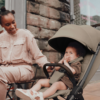 A lifestyle shot featuring a happy mother and her young child using the stroller outdoors. The baby is sitting comfortably in the stroller, while the mother, dressed in neutral tones, smiles at her child. The setting is an urban environment with a brick wall in the background.