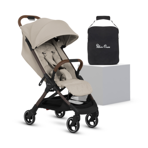 A stylish and compact baby stroller in a light beige "Latte" color, featuring a quilted seat, an adjustable canopy, and a five-point harness. The stroller has a black frame with brown leather-look handles. A black travel bag with the "Silver Cross" logo sits on a white platform beside it.