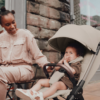 A lifestyle image of a smiling mother interacting with her baby, who is comfortably seated in the beige stroller. The baby is looking playfully at the camera, and the mother, wearing neutral tones, is seated at an outdoor café table with a brick wall in the background.