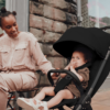 A lifestyle image of a smiling mother interacting with her baby, who is comfortably seated in the black stroller. The baby is looking playfully at the camera, and the mother, wearing neutral tones, is seated at an outdoor café table with a brick wall in the background.