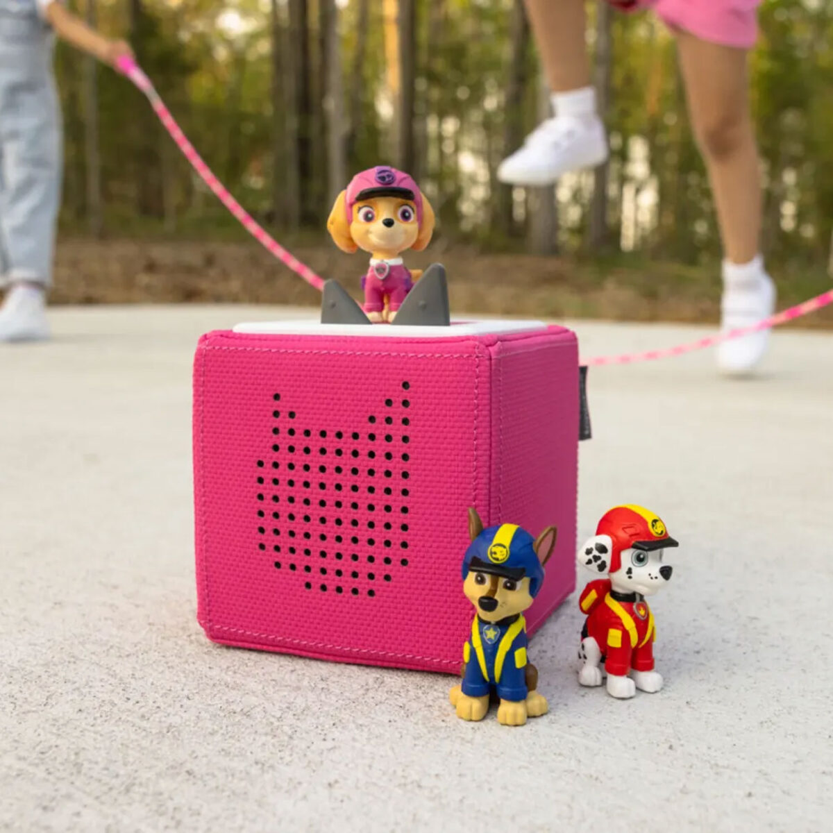 Pink Toniebox with Skye on top and Chase and Marshall beside it, kids playing jump rope in background