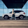 Woman walks past a white SUV carrying the AXKID Up High Back Booster Car Seat in folded mode.