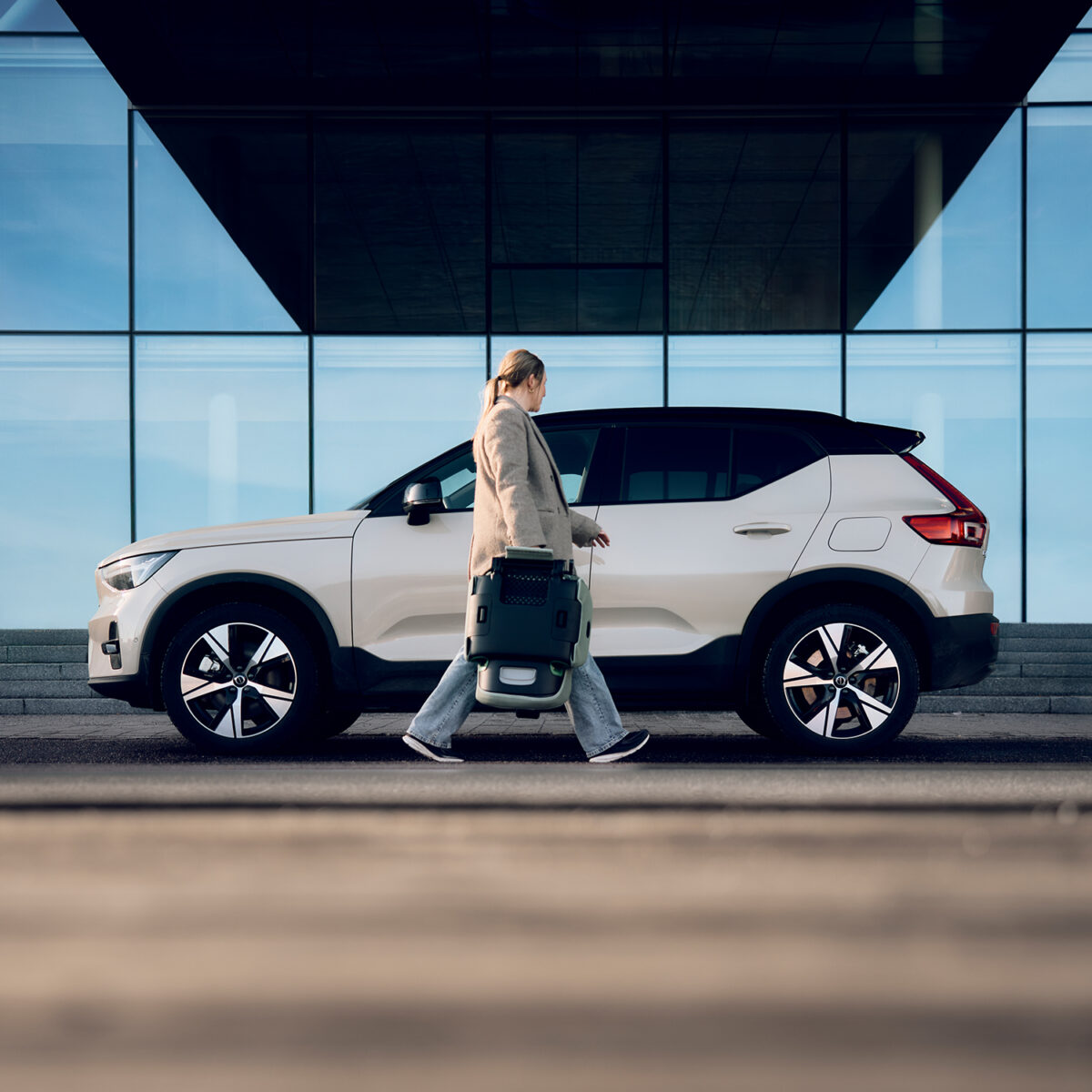 Woman walks past a white SUV carrying the AXKID Up High Back Booster Car Seat in folded mode.