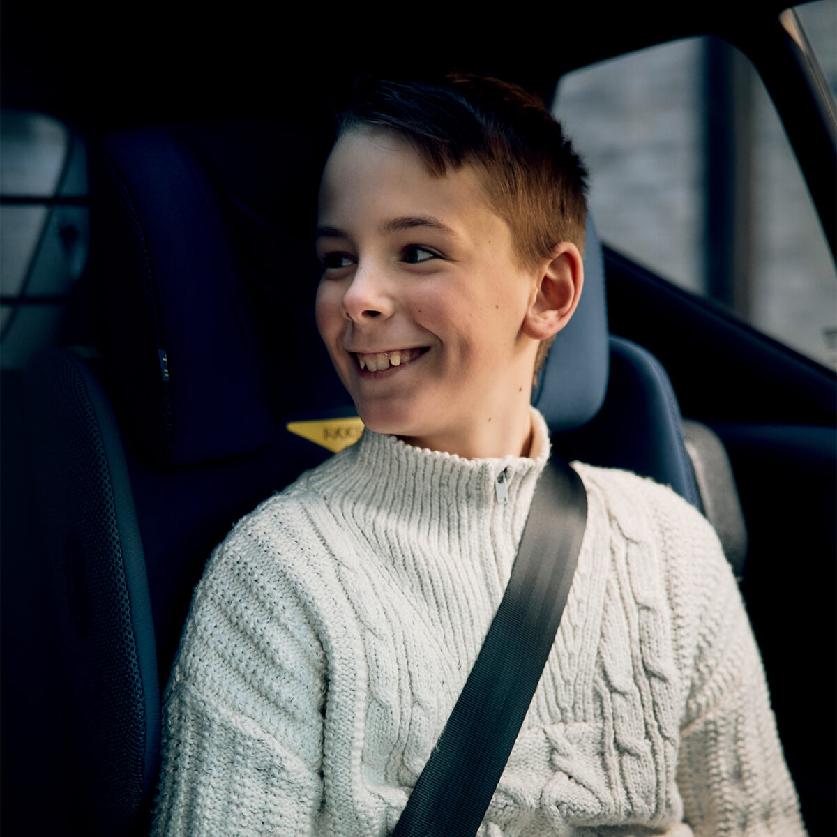 Smiling boy sits safely in the AXKID Up High Back Booster Car Seat wearing a cream knit sweater.