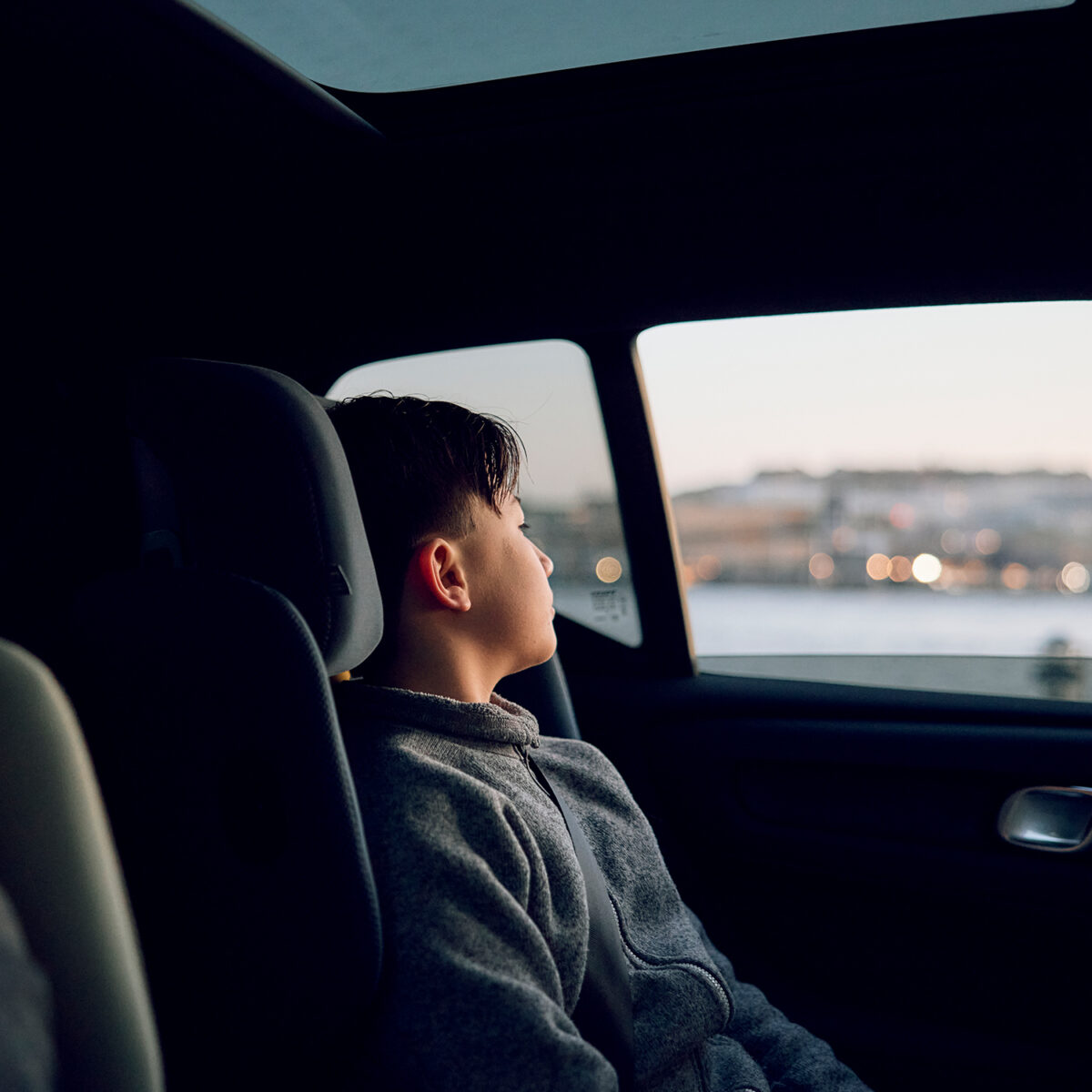 Boy gazing out the window while seated in AXKID Up High Back Booster Car Seat at sunset.