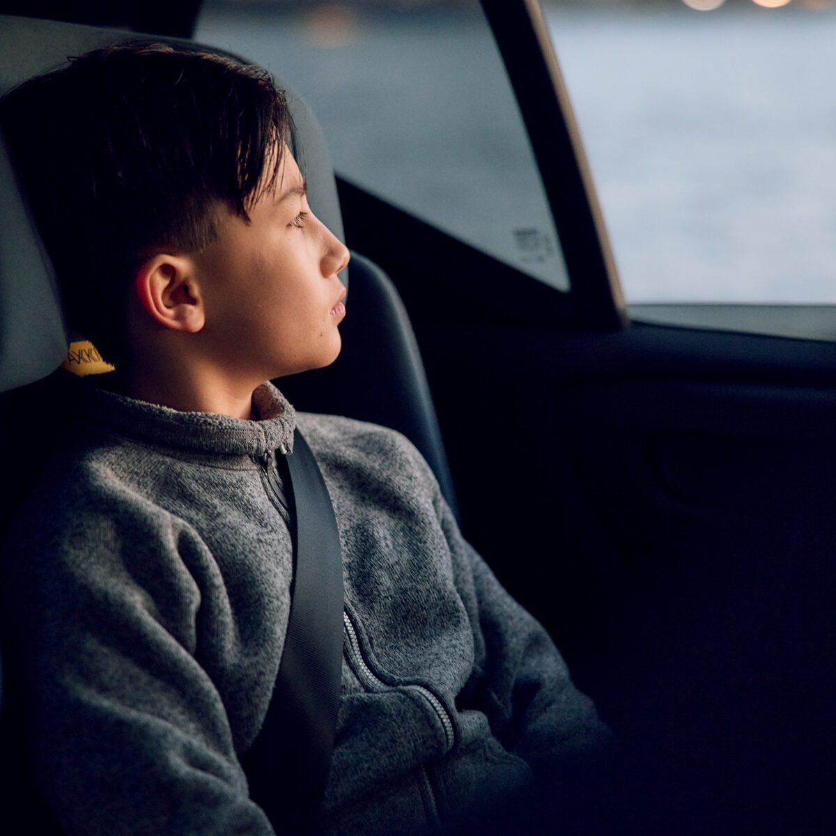 Side profile of a boy seated in AXKID Up High Back Booster Car Seat, looking out a car window.