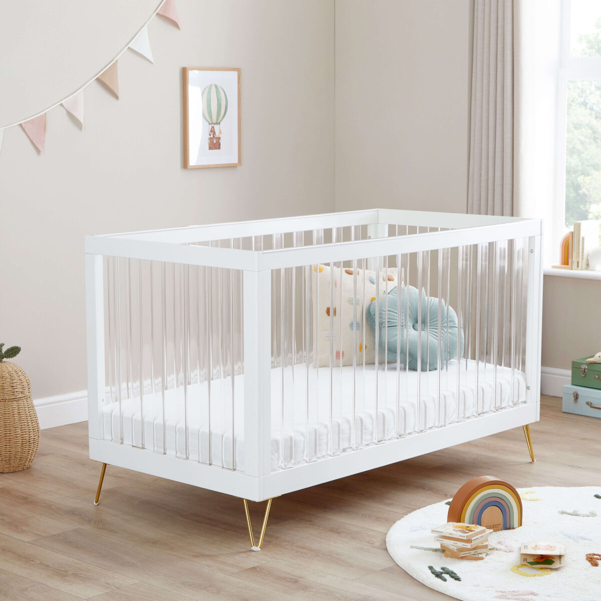 White acrylic cot bed with gold legs, styled in a bright nursery with soft toys and cushions.