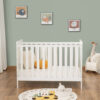 White mini cot bed in nursery with toys, pumpkin pillow and wall art above.