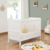White cot bed near window with plush decor, giraffe toy and animal-themed rug.