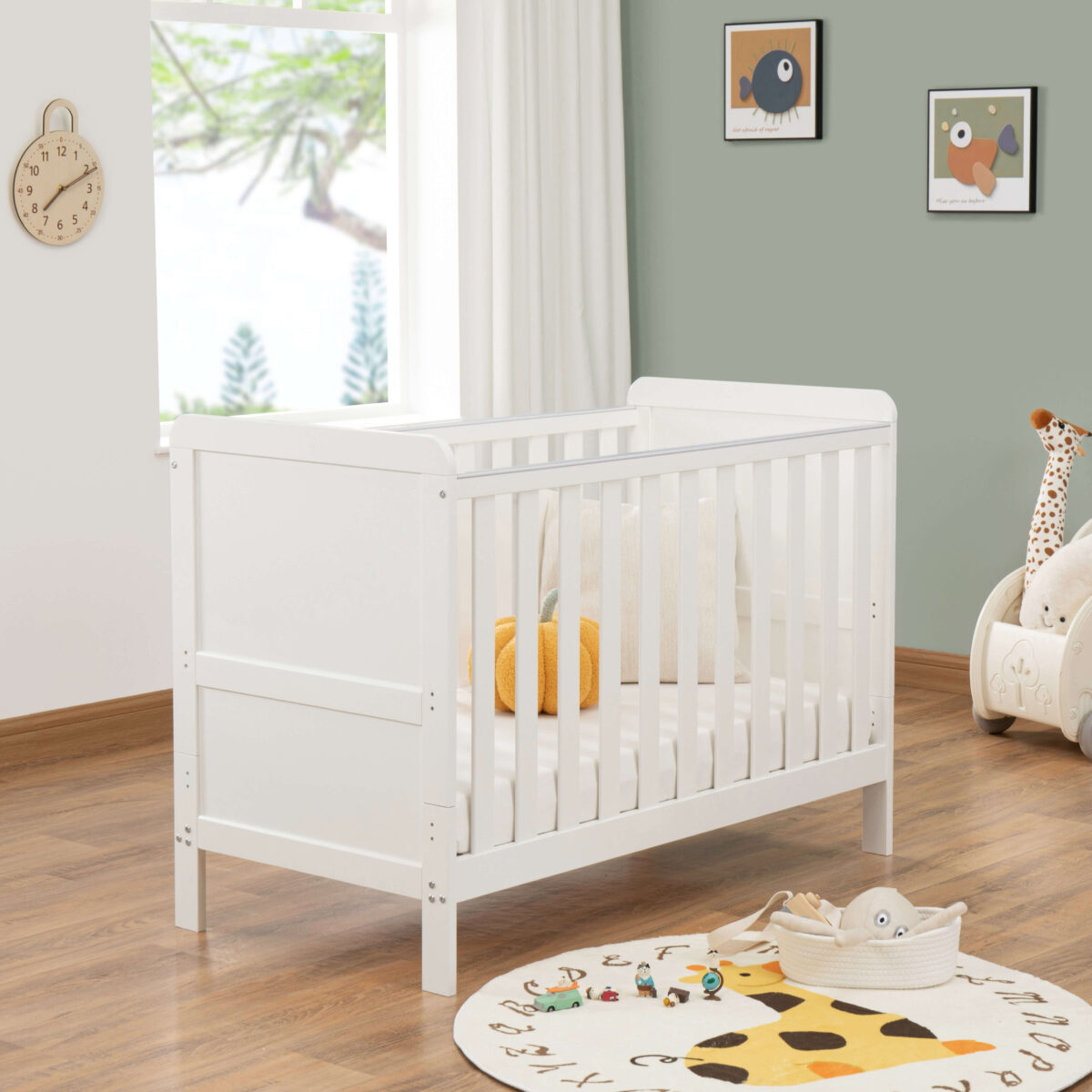 White cot bed near window with plush decor, giraffe toy and animal-themed rug.