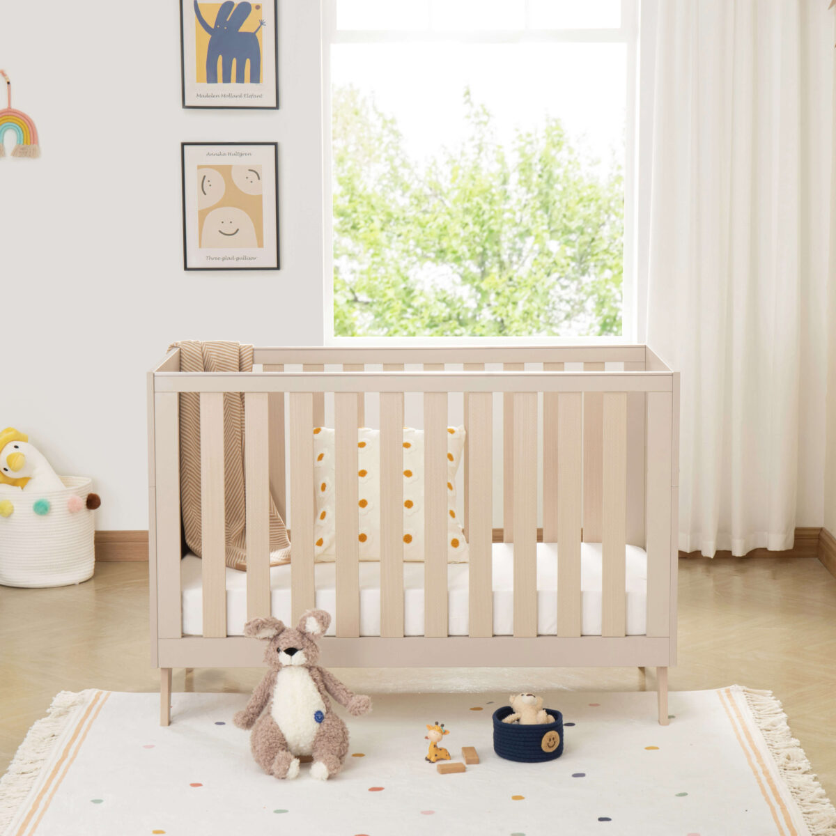 Babymore Dante Mini Cot Bed in cashmere finish with toys and blanket in a soft neutral nursery.