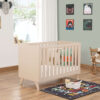 Angled view of Babymore Dante Mini Cot Bed in natural finish, styled in a cosy nursery.