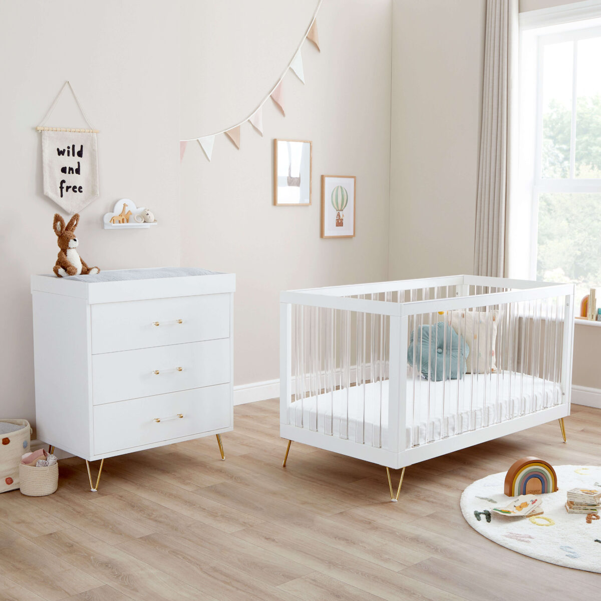 Bright nursery with white crib and dresser, soft toys, rainbow rug, and wall art.