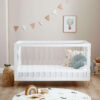 Minimalist white crib with cushions, rainbow toy, and pastel bunting on the wall.